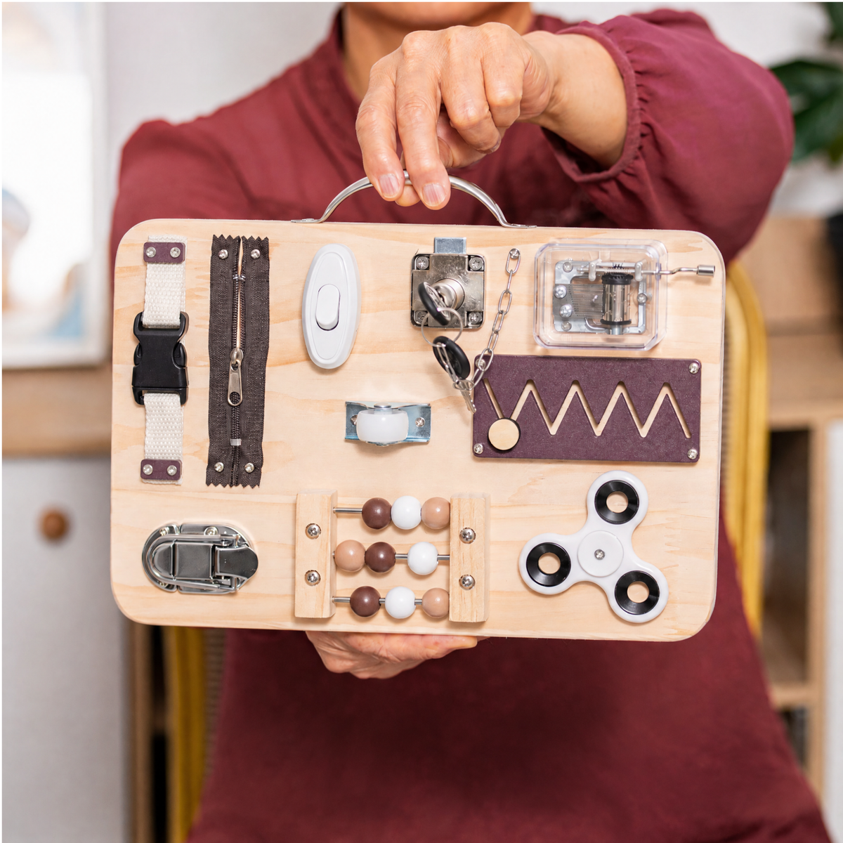 Person holding a wooden busy board by its metal handle, with a buckle, zipper, beads, and spinner — Assistex via EOWD Autism Living