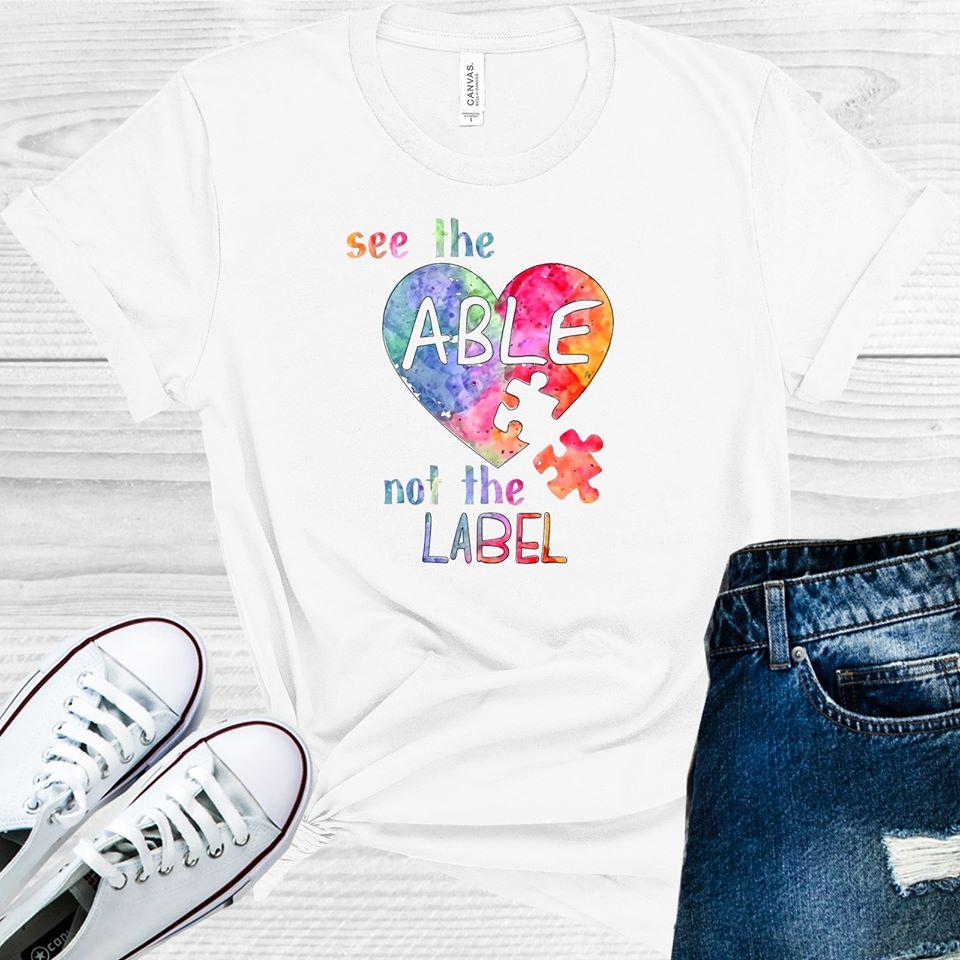 White unisex crew neck graphic tee with colorful heart design and "See the Able Not the Label" text, shown as a flat lay mock-up with sneakers and jeans — Anchored Grace Boutique via EOWD Autism Living